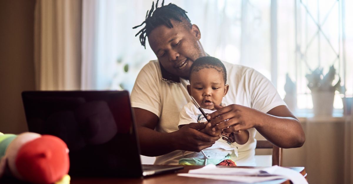 Dad holding his son while working at his desk on a computer from home; why do men hate small groups?